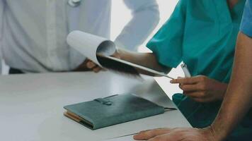 Multiracial medical team having a meeting with doctors in white lab coats and surgical scrubs seated at a table discussing a patients records video