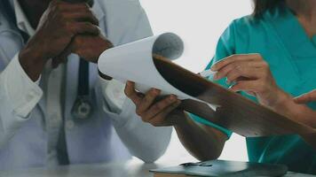Multiracial medical team having a meeting with doctors in white lab coats and surgical scrubs seated at a table discussing a patients records video