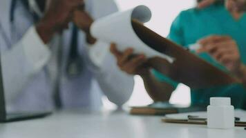 Multiracial medical team having a meeting with doctors in white lab coats and surgical scrubs seated at a table discussing a patients records video