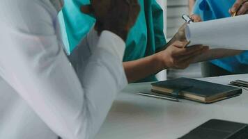 Multiracial medical team having a meeting with doctors in white lab coats and surgical scrubs seated at a table discussing a patients records video