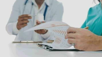 Multiracial medical team having a meeting with doctors in white lab coats and surgical scrubs seated at a table discussing a patients records video