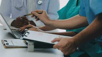 Multiracial medical team having a meeting with doctors in white lab coats and surgical scrubs seated at a table discussing a patients records video