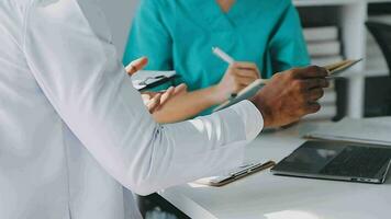 Multiracial medical team having a meeting with doctors in white lab coats and surgical scrubs seated at a table discussing a patients records video