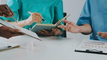 Multiracial medical team having a meeting with doctors in white lab coats and surgical scrubs seated at a table discussing a patients records video