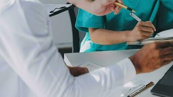 Multiracial medical team having a meeting with doctors in white lab coats and surgical scrubs seated at a table discussing a patients records video