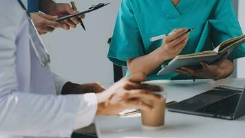 Multiracial medical team having a meeting with doctors in white lab coats and surgical scrubs seated at a table discussing a patients records video