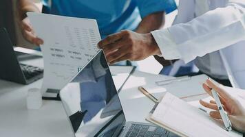 Multiracial medical team having a meeting with doctors in white lab coats and surgical scrubs seated at a table discussing a patients records video