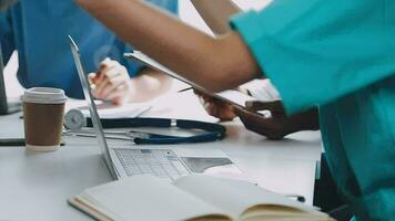 Multiracial medical team having a meeting with doctors in white lab coats and surgical scrubs seated at a table discussing a patients records video