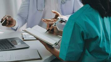 Multiracial medical team having a meeting with doctors in white lab coats and surgical scrubs seated at a table discussing a patients records video