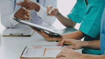 Multiracial medical team having a meeting with doctors in white lab coats and surgical scrubs seated at a table discussing a patients records video
