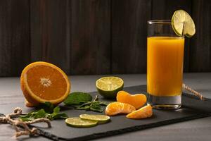 Orange juice in a glass with a slice of lime on a black board on a dark wooden background. Near orange, mint, tangerine slices. Healthy food photo