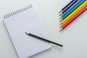 Clean notepad in a cage with a pencil lying on it and color pencils in the form of an arrow on a white background top view photo