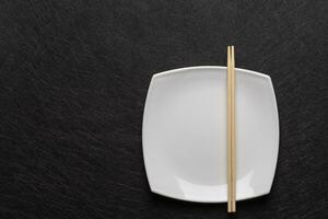 vacío blanco plato con palillos en un oscuro antecedentes. japonés comida estilo. parte superior ver plano laico con Copiar espacio foto