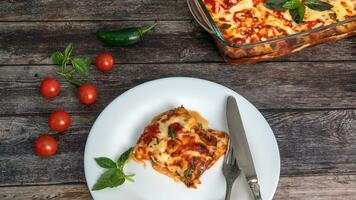 Eggplant parmigiano, a piece on a plate with a knife and fork, next to the tray with eggplants, cherry tomatoes photo