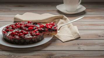 Pie with berries, raspberries, strawberries, currants, on a white plate, next to a piece of cake on the shoulder. On a wooden background, in the background napkin, white cup photo