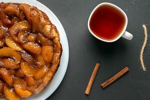 tarta tatin con duraznos y caramelo en un blanco plato en un oscuro antecedentes. siguiente a el blanco taza con negro té y canela palos foto