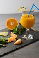 Orange juice in a glass with tube on a black board on a light background. Near orange, mint, tangerine slices. Healthy food photo