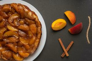 Tarte Tatin with peaches and caramel on a white plate on a dark background. Near slices of peach and cinnamon sticks. Top view, horizontal orientation photo