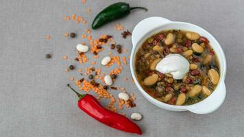 Mexican soup of seven kinds of beans with sour cream, closeup, on a gray linen background surrounded by red pepper and beans photo