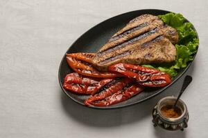 Grilled Turkey breast steak with baked peppers and lettuce leaves in a black plate on a light linen background. Next to the pepper shaker with red pepper. Horizontal orientation, copy space photo