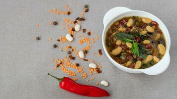 Mexican soup of seven kinds of beans with basil, closeup, on a gray linen background surrounded by red pepper and beans photo