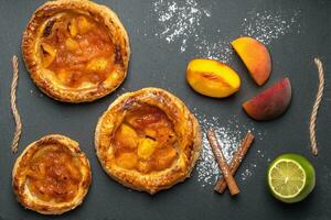 Homemade round puffs with peaches on a dark background, next to peach slices, cinnamon sticks, lime, powdered sugar. Top view, horizontal orientation photo