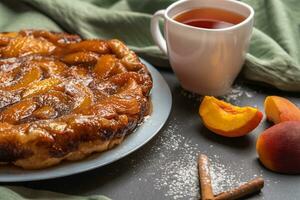 arte tatin con melocotones, caramelo y en polvo azúcar de cerca en un blanco plato en un oscuro antecedentes con un lino servilleta. siguiente a el blanco taza con negro té, melocotón rebanadas y canela palos foto