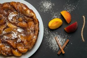 Tarte Tatin with peaches, caramel and powdered sugar on a white plate on a dark background. Near slices of peach and cinnamon sticks. Top view, horizontal orientation photo