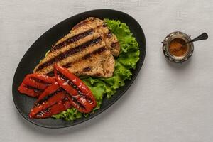 Grilled turkey breast steak with baked peppers and lettuce leaves in a black plate on a light linen background. Next to the pepper shaker with red pepper. Top view, copy space photo