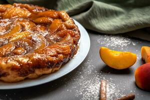 Tarte Tatin with peaches, caramel and powdered sugar closeup on a white plate on a dark background with a linen napkin. Near slices of peach and cinnamon sticks. Horizontal orientation photo