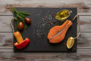 Raw salmon steak. On a dark board. Near baked peppers, tomatoes, lemon. Olive oil in an oil can. Healthy food. Top view photo