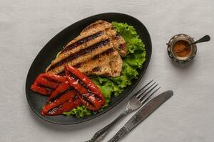 Grilled turkey breast steak with baked peppers and lettuce leaves in a oval black plate with knife and fork on a light linen background. Next to the pepper shaker with red pepper. Top view, copy space photo