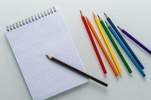 Clean notepad in a cage with a pencil lying on it and rainbow color pencils on a white background top view photo