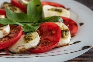 blanco plato de sano clásico delicioso caprese ensalada con Tomates y queso Mozzarella queso con albahaca hojas y tiras de soja salsa en el madera antecedentes. cerca arriba. foto