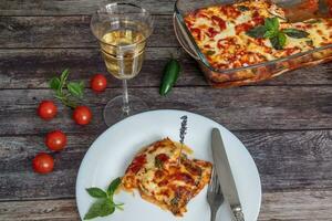 Eggplant Parmigiano, a piece on a plate with a knife and fork, next to the tray with eggplants, a glass of white wine, cherry tomatoes photo