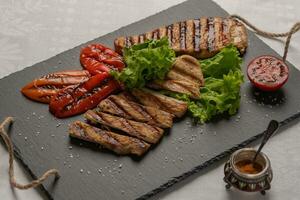 Grilled turkey breast sliced steaks with baked peppers,lettuce and tomato on a black board. Next to the pepper shaker with red pepper. Horizontal orientation, closeup photo