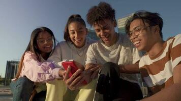 Group of happy teenage friends looking the phone and laughing in a bench in the city street. video