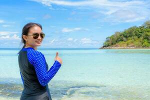 contento asiático mujer torneado a sonrisa con pulgar arriba a el playa foto