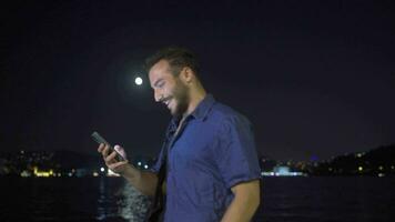 jung Mann mit Telefon beim Nacht gegen Stadt Aussicht durch das Meer. video