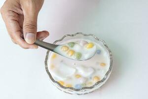 Hand holding spoon to scooping rice balls with young coconut in sweet coconut milk in a cup isolated on white  tile background Thai people call Bua Loi. photo