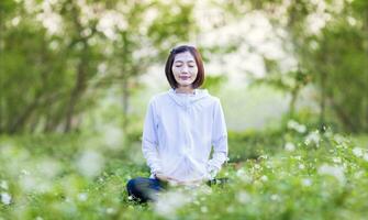 asiático mujer es relajantemente practicando meditación yoga en el bosque lleno de margarita flor en verano a alcanzar felicidad desde interior paz sabiduría con Mañana ligero para sano mente y alma concepto foto