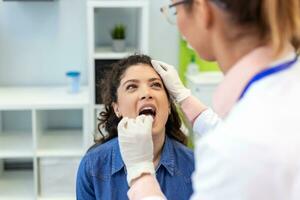 un joven mujer se sienta en un examen mesa a través de desde su doctor. el médico alcanza adelante con un lengua depresor como el mujer mira arriba y palos fuera su lengua. foto