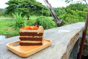 A piece of chocolate layer cake with topping macadamia nut with caramel on light brown wood plate on old plank in natural environment, Thailand. photo