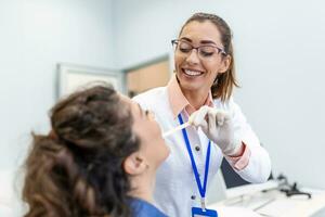 A young woman sits on an exam table across from her doctor. The doctor reaches forward with a tongue depressor as the woman looks up and sticks out her tongue. photo