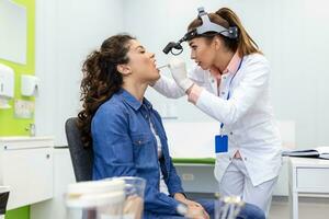 Female patient opening her mouth for the doctor to look in her throat. Female doctor examining sore throat of patient in clinic. Otolaryngologist examines sore throat of patient. photo