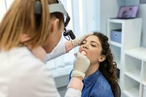 Female patient opening her mouth for the doctor to look in her throat. Female doctor examining sore throat of patient in clinic. Otolaryngologist examines sore throat of patient. photo