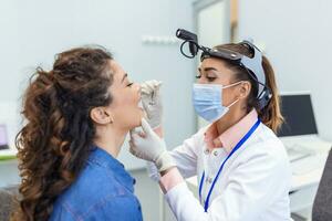 A young woman sits on an exam table across from her doctor. The doctor reaches forward with a tongue depressor as the woman looks up and sticks out her tongue. photo