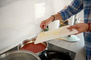 de cerca de del chef manos poniendo Cortado ajo desde un corte tablero dentro un cacerola con hirviendo tomate salsa foto