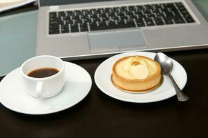 It's time to take a coffee break. A cup of freshly brewed espresso and sweet dessert near a laptop. photo
