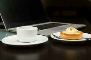 Still life with food and electronical device. A sweet delicious tartlet, cup of coffee over laptop background. photo